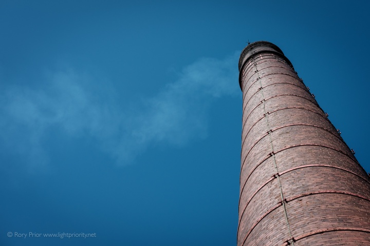 Looking up at a lightly smoking, red brick mill chimney with a cloudless blue sky behind.